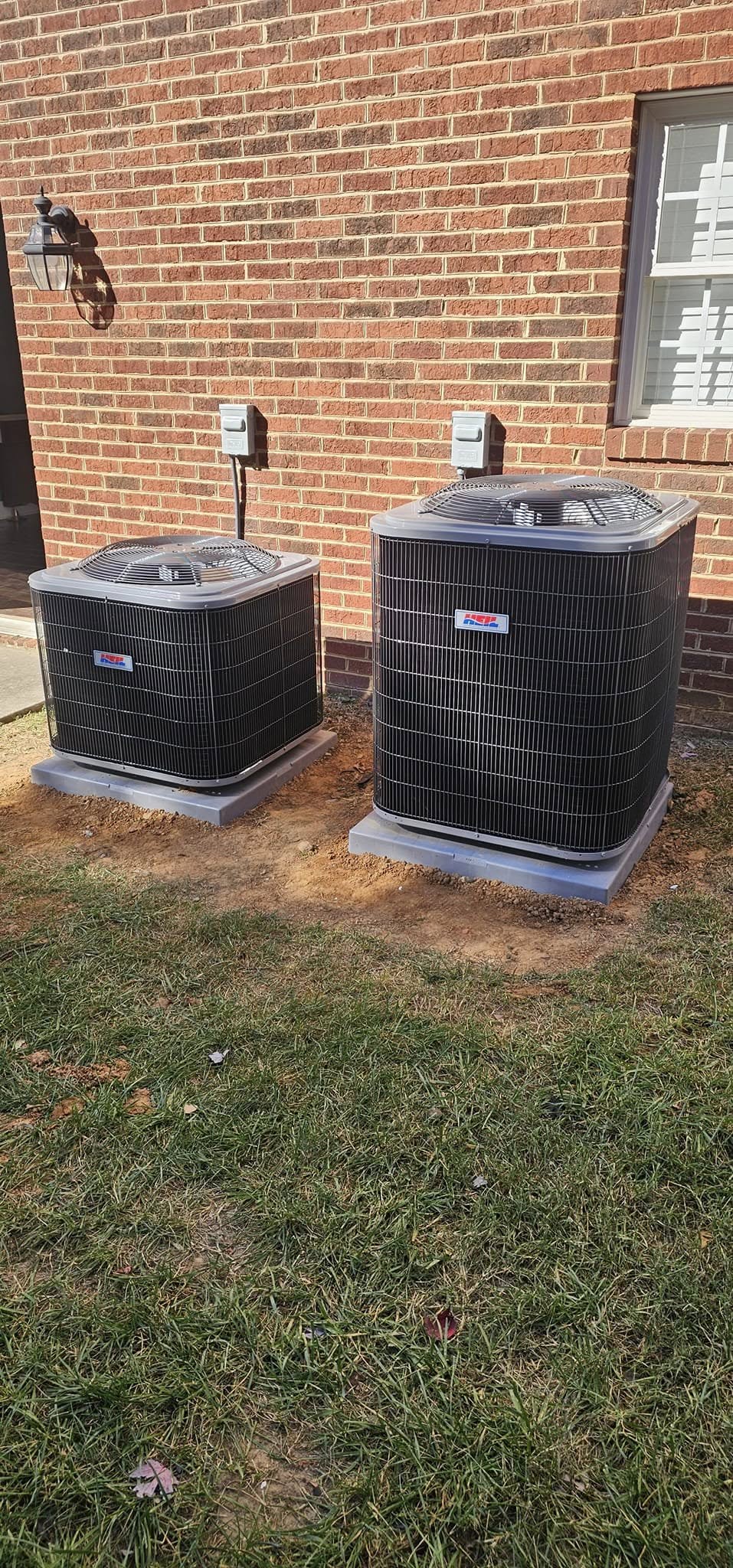 Two outdoor air conditioning units are installed on concrete pads next to a brick house, with some grass and soil visible in the foreground.