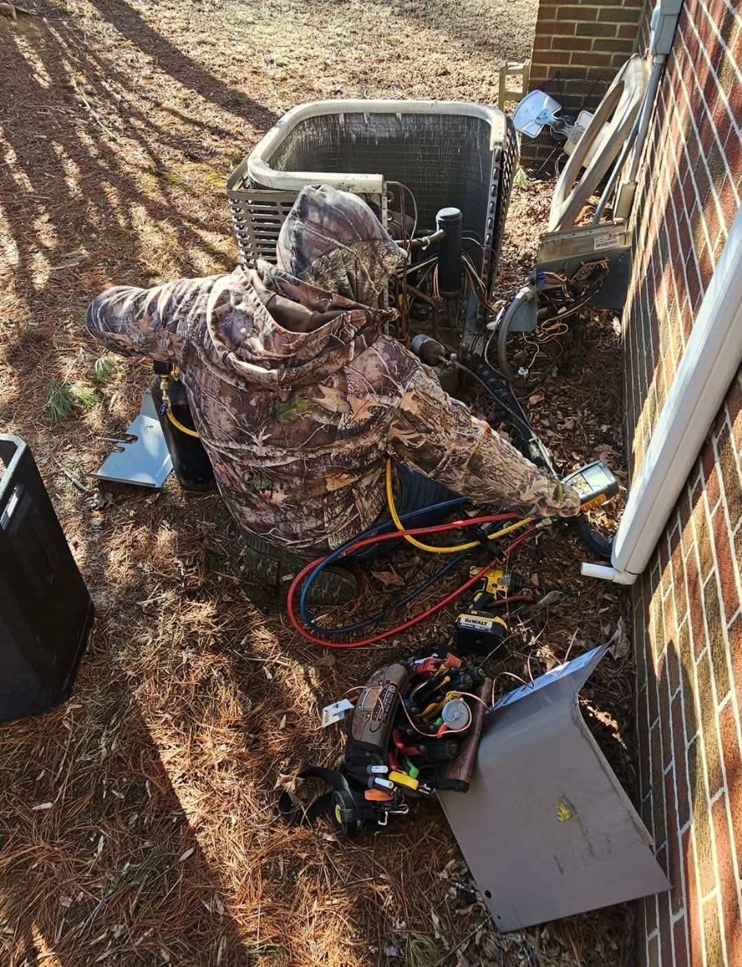 A person in camouflage clothing repairs an outdoor HVAC unit next to a brick wall, with tools and equipment spread on the ground nearby.