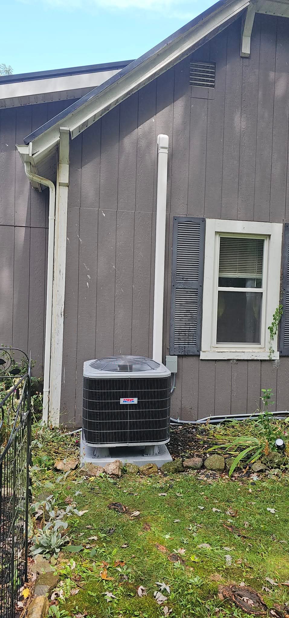 An air conditioning unit is installed outside a gray house with white trim and black shutters, next to a window and a small garden area.