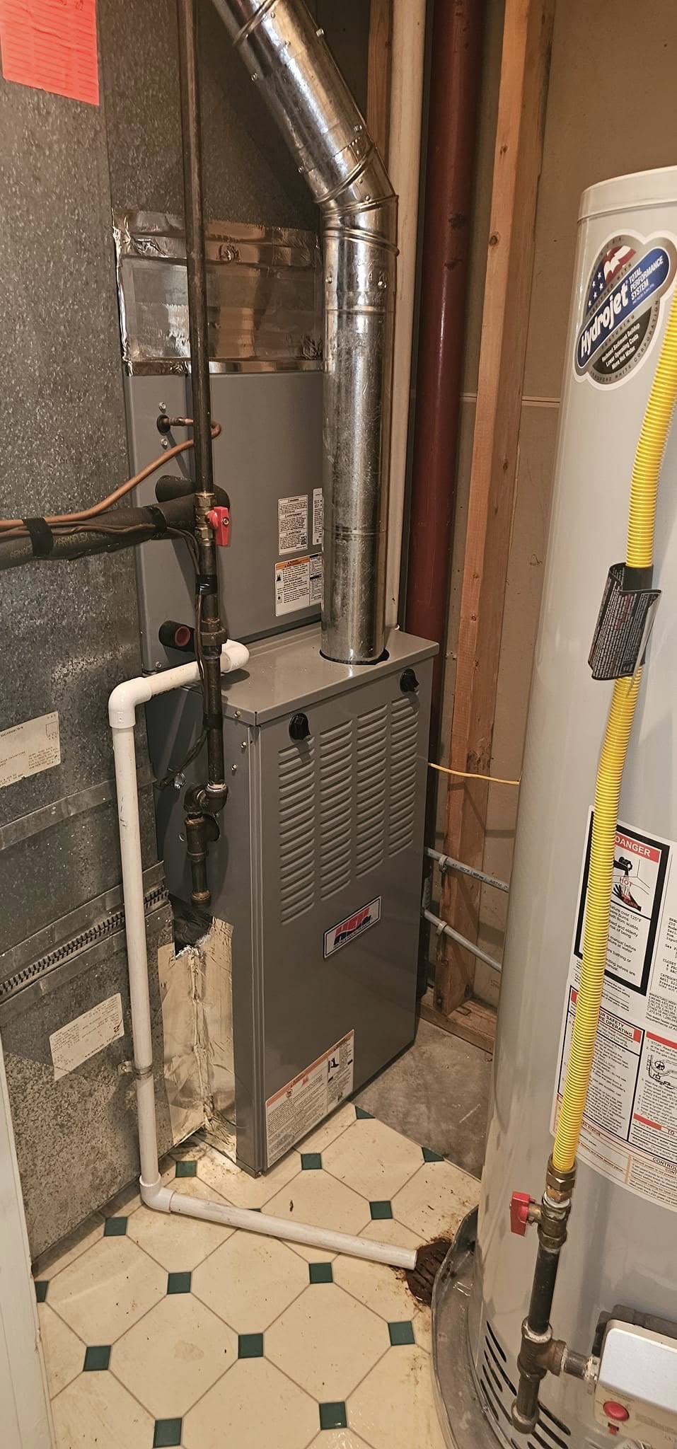 A gray furnace unit connected to metal ductwork and white PVC pipes is installed next to a water heater with a yellow gas line in a utility room with exposed walls.