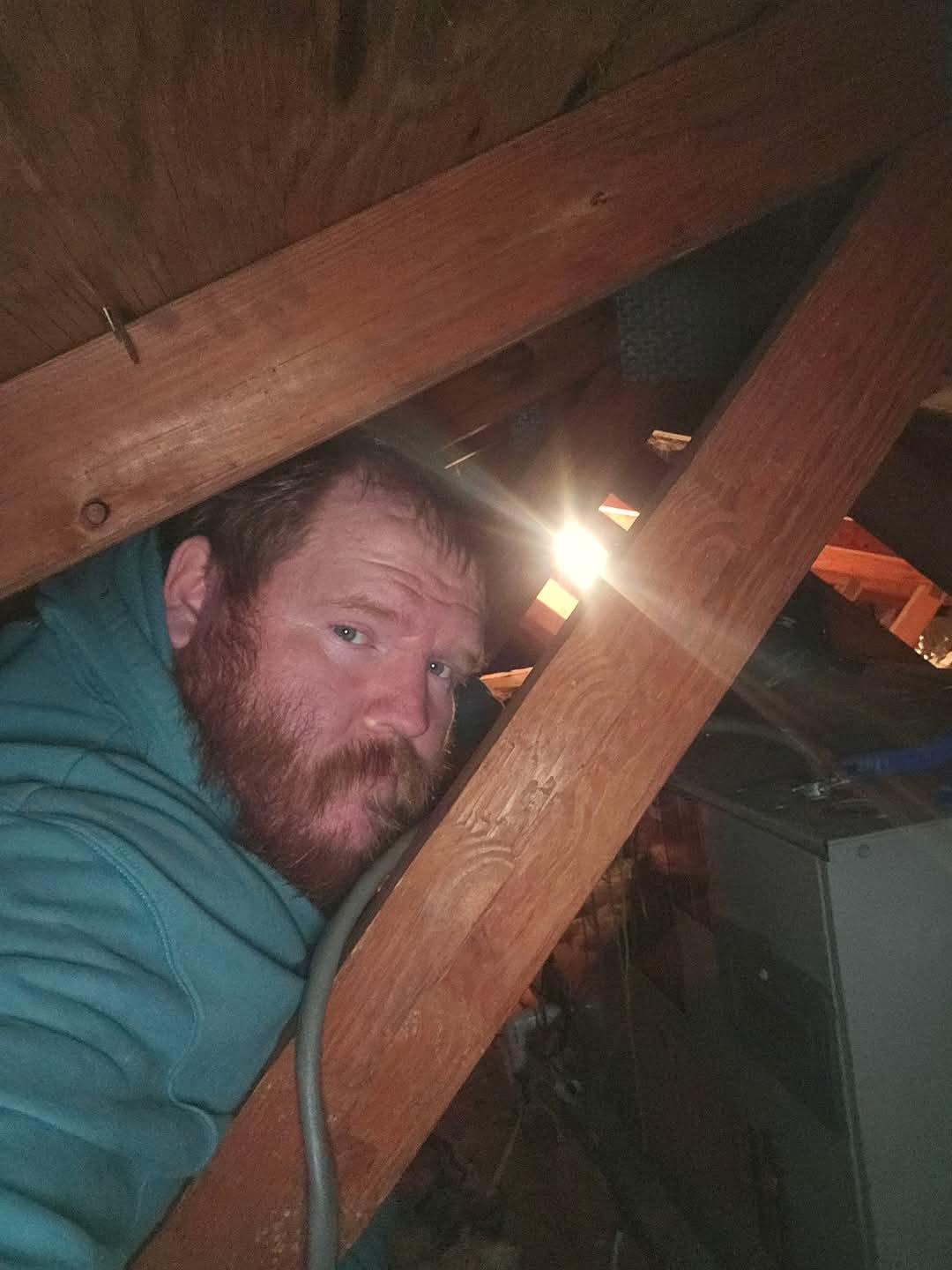 A man in a teal hoodie sits in an attic space next to wooden beams and HVAC equipment, with a bright light shining behind him.
