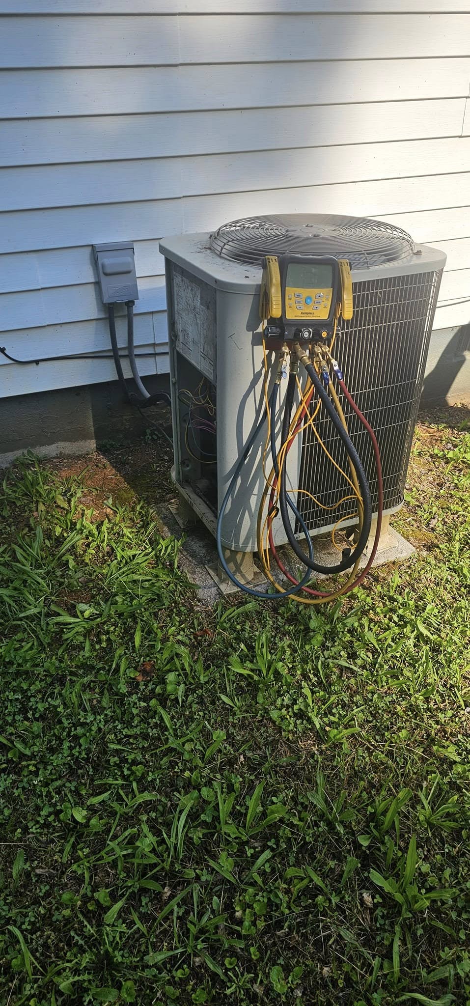 An air conditioning unit is positioned outside a house, connected to gauges and hoses for maintenance or inspection.