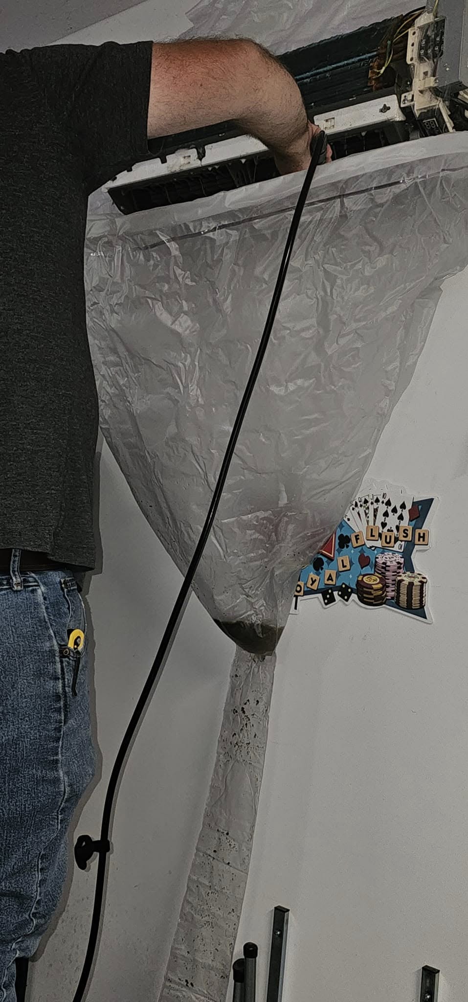A person is cleaning an air conditioning unit using a hose, with a plastic sheet attached below to catch debris or water.