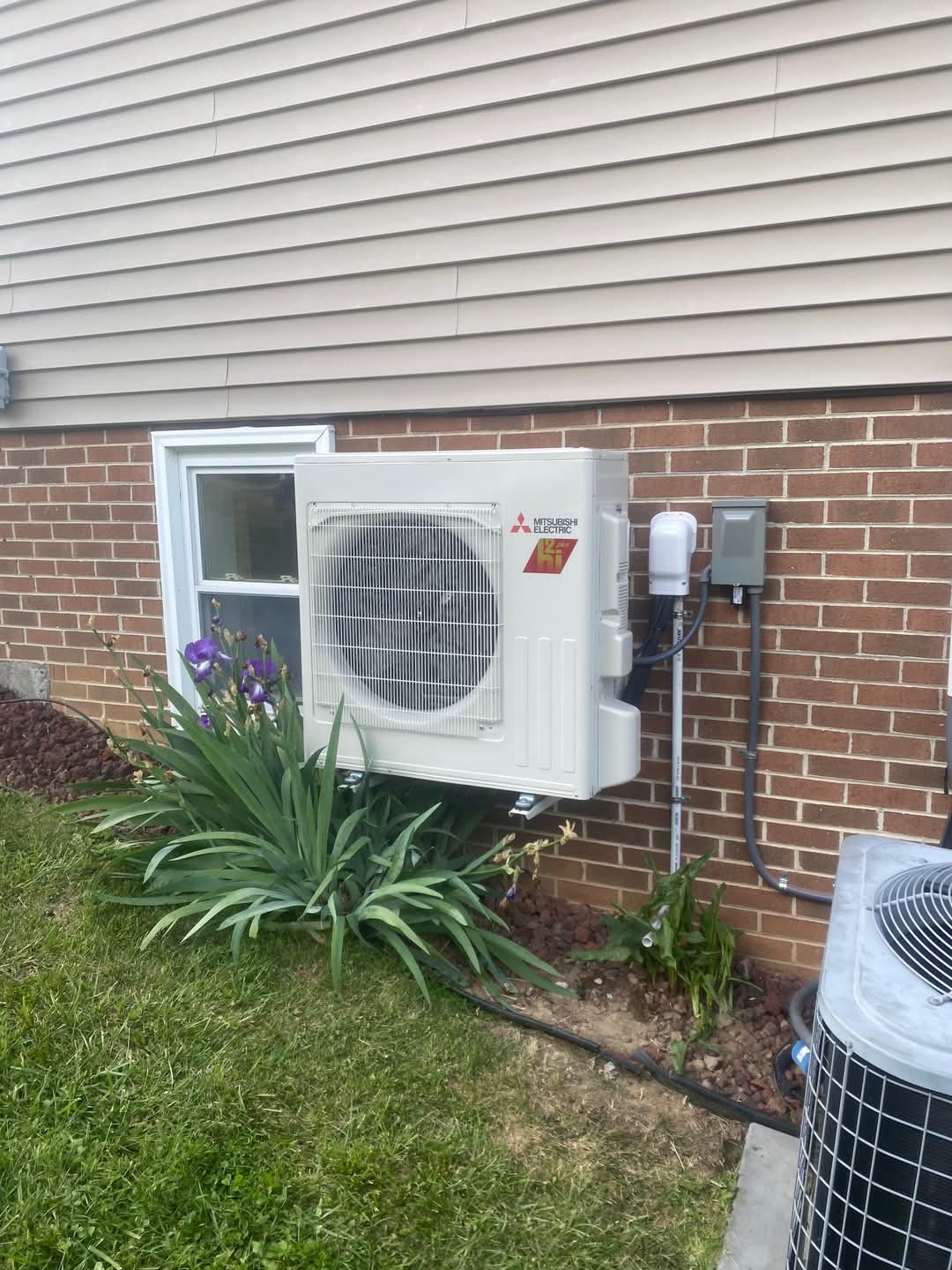 An outdoor Mitsubishi air conditioning unit is mounted on the exterior wall of a brick house above some green plants near a small window.