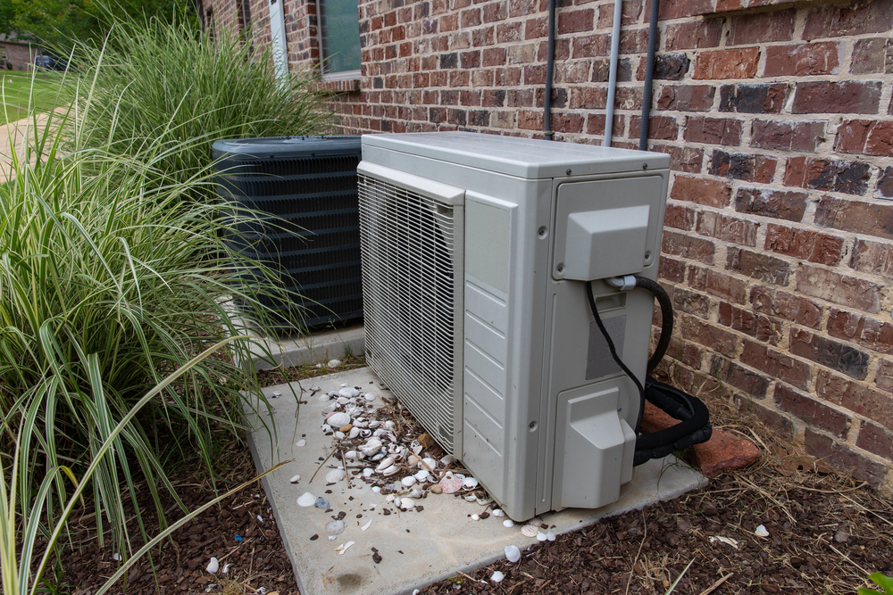 Two outdoor air conditioning units sit on concrete pads next to a brick wall, surrounded by ornamental grass and scattered debris.